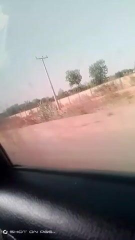The video depicts a view from inside a moving vehicle, likely a car. The camera is pointed towards the passenger side window, capturing the passing scenery. The landscape consists primarily of a dry, dusty environment with sparse vegetation. A dirt road or path runs parallel to the vehicle's trajectory, with a low fence or wall visible along the roadside. Utility poles are sporadically placed along the road. The sky appears hazy and light-colored. The interior of the vehicle is visible at the bottom of the frame. There are no people distinctly visible in the video. The overall impression is that the video was recorded in a rural or undeveloped area.