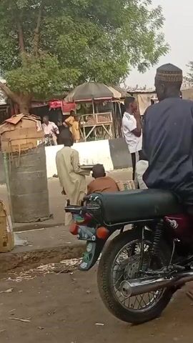 The video captures a scene in a bustling outdoor environment. A man sits on a maroon motorcycle wearing a dark shirt and a patterned cap. Around him are other individuals, some dressed in traditional clothing, and others in modern attire. To the left, watermelons are displayed on a concrete platform. There is a modest structure labeled "EDUCATIONAL MATERIALS AND SHOES". People are standing, talking, and generally interacting in this busy setting. The video is shot at ground level, slightly angled to include as much of the active scene as possible.