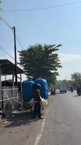 A street vendor stands next to a cart filled with goods, including large blue bags and what appear to be bags of snacks. The vendor is wearing a cap, a jacket, and jeans. The cart is parked on the side of a paved road. In the background, there are trees, buildings, a utility pole, and vehicles moving on the road. The sky is clear and blue. The overall scene depicts a typical street market environment on a sunny day.
