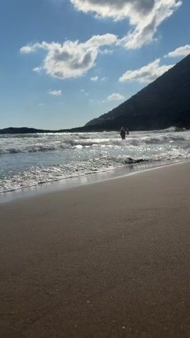 The video shows a beach with sandy shore in the foreground. The ocean waves are breaking on the shore. In the background, there are a few people in the water. A hill covered with vegetation is visible on the right side of the frame. The sky is blue with some white clouds. The sun is shining. The video is shot from a low angle, close to the ground.