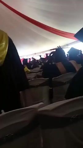 The video shows a graduation ceremony taking place inside a large tent. The tent has a white ceiling with red stripes. Rows of chairs covered in white fabric are set up for the audience. People wearing graduation gowns and caps are walking around, some with stoles of different colors. The camera appears to be held by someone sitting among the audience, moving slightly.