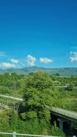 The video captures a landscape scene from a moving vehicle, likely a train or car, looking out a window. The sky is a clear, vibrant blue with a few scattered white clouds. Below the sky, rolling green hills are visible, dotted with trees and some buildings. In the foreground, there is a vineyard with rows of plants supported by trellises. The perspective shifts slightly as the vehicle moves, giving a sense of travel through the countryside. The overall impression is one of a sunny day in a rural, possibly agricultural, area. The audio is a person speaking Ukrainian, but the content is unintelligible in this context.