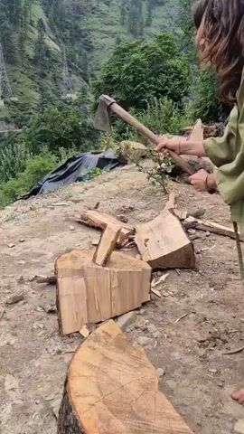 The video captures a person in Shila, India, engaged in the activity of chopping firewood. The scene is set outdoors on a dirt ground, with a lush green mountainside covered in trees and vegetation in the background. The weather appears to be overcast or hazy, suggesting it might be a cloudy day or early morning.

The primary subject is a person, whose gender is not definitively clear but they have long, dark hair. They are wearing a light green garment and are barefoot. The person is actively splitting logs of wood with an axe. Several pieces of chopped wood are scattered around them, indicating that this is an ongoing task. The logs themselves appear to be of a substantial size, suggesting they will provide a good amount of firewood.

In the background, there are some electrical pylons visible on the mountainside, hinting at the presence of infrastructure in the otherwise natural setting. A large piece of black plastic sheeting is also visible near the chopped wood, possibly used to cover other materials or as part of the preparation for storing the firewood.

The focus of the video is on the physical labor of chopping wood. The person is seen swinging the axe with force and precision. The overall mood of the video is one of practical work and self-sufficiency in a rural environment. No text is visible in the video.