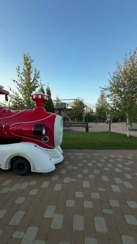 A red and white toy train with a driver and passengers moves slowly along a paved path. The train has multiple cars, each with windows and doors. In the background, there is a grassy area with trees and a clear blue sky.