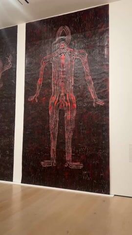 The video shows the interior of an art gallery. The room has light wooden floors, white walls, and a section of deep red wall. There are recessed lighting fixtures in the ceiling. On the white walls are two large, dark paintings that depict figures outlined in red. In the distance, near the red wall, is a structure constructed from light-colored branches. A person wearing a black outfit is walking through the gallery. The camera seems to be moving through the gallery as well, panning from the paintings to the other objects and the person.
