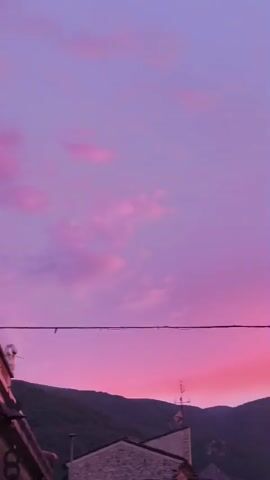 The video shows a time-lapse of a sunset over a mountain range. The sky transitions from a pale purple to a vibrant pink and red as the sun sets. In the foreground, there are rooftops of houses and trees silhouetted against the colorful sky. A power line is visible across the frame. The video captures the changing colors of the sky and the deepening shadows as evening approaches.
