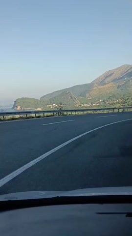 The video shows a view from the inside of a vehicle, looking out at a highway with mountains in the background. The highway has two lanes in the direction of travel, marked with white lines. A guardrail runs along the side of the road, separating it from a steep, tree-covered mountainside. The sky is clear and blue, with the sun casting shadows on the mountains. The mountains are rugged and appear to be sparsely vegetated in some areas, with denser green foliage in others. The vehicle appears to be moving at a moderate speed.