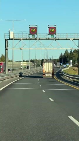 The video shows a view from a moving vehicle on a highway. The road is paved and has clear lane markings. Overhead gantries display speed limits of 130 with downward arrows. A large white truck with a trailer is visible in front, traveling in the same direction. Other vehicles are present in the distance on the highway. The weather appears clear with a blue sky, and trees and foliage line the side of the road. Streetlights are also visible along the highway.
