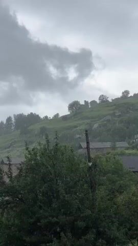 The video displays a gray, overcast sky with heavy clouds. Rain is falling, visible as streaks against the backdrop of a lush green hillside. Several trees are in the foreground, some with dark green foliage. The hillside is dotted with scattered houses and some scattered trees. A utility pole stands to the right of the frame. The scene evokes a rural, possibly mountainous, environment during a rain shower.