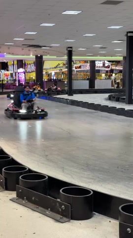 Several individuals are captured in a video, actively participating in an indoor go-kart race. They are seen navigating a smooth, light grey track that features black cylindrical barriers along its edges. One participant, wearing a blue shirt, is prominently featured driving a black go-kart. Other go-karts, some equipped with blue underglow lights, follow closely. In the brightly lit background, an arcade area is visible, adorned with various games, flashing lights, and signs, including one that clearly displays the word "NUCLEAR." The setting appears to be a large entertainment complex, possibly in Columbus, United States of America, with artificial lighting illuminating the entire space.