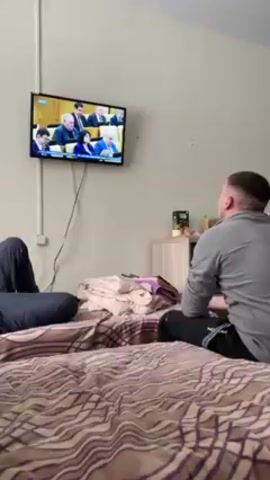 Two men are on a bed in what appears to be a simple room. One man is lying down, facing away from the camera. The other man sits cross-legged and faces a wall-mounted television, which displays an image of several people, including Vladimir Putin. A small bedside table sits near the head of the bed, with some items placed on top. The room is sparsely decorated.