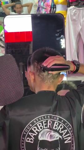 A barber is cutting a customer’s hair in what appears to be an outdoor market setting. The barber, wearing a black jacket with the words “BARBER BRAIN” and a logo of a head with stylized hair, is using a black hair clipper on the back of the customer’s head. The customer is facing a mirror, and the barber uses a handheld mirror for the customer to view the progress. The background shows clothing items hanging on racks and other people in the vicinity. The lighting is uneven.