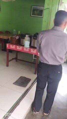 The video begins with a rear view of a man wearing a grey collared shirt and black pants, walking away from the camera down a narrow corridor. He is barefoot. The corridor appears to be an outdoor walkway. To the left of the corridor, there is a room with a table covered in a pink tablecloth, a large bottle, and two dispensers. There is also a framed picture on the wall. To the right of the corridor, the walkway opens into a room with several red plastic chairs arranged around tables. In the background of this room, a woman wearing a pink hijab and a light brown top with purple pants is visible. She is barefoot and walks towards the camera. The room appears to be a dining area. The walls are painted green. The floor is tiled. The video pans slightly to the right, showing more of the dining area. The woman continues to walk, and the video ends as she moves further into the room.