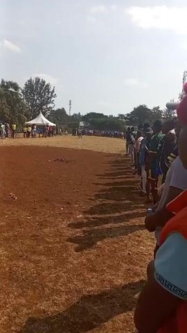 A crowd of people stands along a dirt field watching a person run toward the finish line. The field is reddish-brown, and the sky is bright. A tent is visible in the distance. The runner is wearing a green shirt.