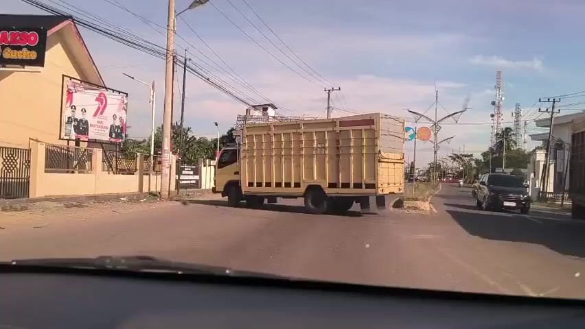 The view from inside a vehicle shows a two-lane road with light traffic in a small town. A yellow truck with a gray-covered cargo area is making a left turn at an intersection. A motorcycle rider wearing a white top and helmet passes the truck on the left. A black car is approaching from the opposite direction in the right lane. Power lines and poles run along the street. Buildings line both sides of the road.  A billboard stands on the left side of the road at the edge of a fenced property. The sky is blue with few clouds.