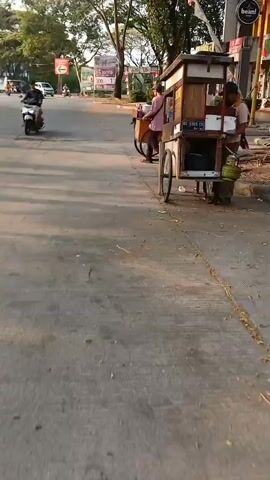 A street vendor pushes a food cart down a paved road. The cart is wooden with two large wheels and a smaller wheel in front. It has a glass display case for food and a propane tank on the side. A man in a light pink shirt and dark pants is seen at the cart, possibly serving a customer. Another person in a light pink shirt is walking away from the cart in the background. The street appears to be in an urban or suburban area, with buildings and trees visible on the side. There are also motorcycles parked on the side of the road. The lighting suggests it is either early morning or late afternoon.