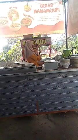 The video opens with a close-up of a food stall with a banner advertising "BAKSO PRAZMANZAN". A woman wearing an orange hijab and outfit is visible behind the counter. The counter itself is covered with a grey, wood-patterned material. To the right of the woman, there are stacks of bowls and plates, as well as some containers. The camera pans to the right, revealing more of the stall's interior, including a white refrigerator. The scene then shifts to a different perspective, showing a table with a red and white checkered tablecloth. On the table, there is a purple lidded container, a yellow bowl with a silver can inside, and some red utensils. A plastic bottle is hanging from above. In the background, there is a street with trees and some buildings. The video concludes with a close-up of an orange control panel with red and green buttons, and a red switch.