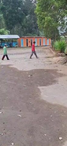 Two men are walking in an outdoor area. One man is wearing a green shirt and black pants, and the other man is wearing a red shirt and grey pants. They are walking towards a building that has several orange doors with blue trim. There are trees and foliage in the background, and the ground is dirt with some puddles of water.