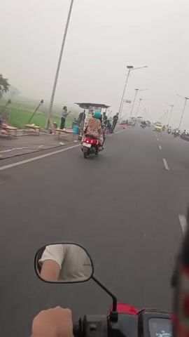The video shows a point-of-view shot from a moving motorcycle on a hazy road. In the foreground, a hand grips the motorcycle's handlebar. A side mirror reflects the rider's arm and shoulder. On the side of the road, there are several stalls set up, with vendors and customers. Some people are standing, while others are seated. There are also motorcycles and a few cars on the road, suggesting traffic. In the background, there are rice fields and tall streetlights with solar panels. The overall atmosphere is slightly obscured by a thick haze, possibly due to weather or pollution.