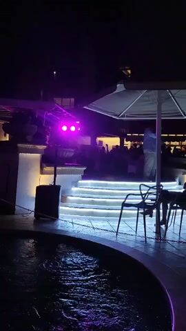 The video captures an outdoor scene at night, illuminated by purple and white lights. In the foreground, a circular pool of water reflects the purple light. Beyond the pool are illuminated steps leading up to an area with tables and chairs, shaded by a large umbrella. There are several people visible in the background, some seated at tables, and one standing near the top of the stairs. A speaker is positioned to the left of the stairs, and a lighting rig with purple lights is mounted on a stand. The overall atmosphere appears to be a lively outdoor venue with ambient lighting.