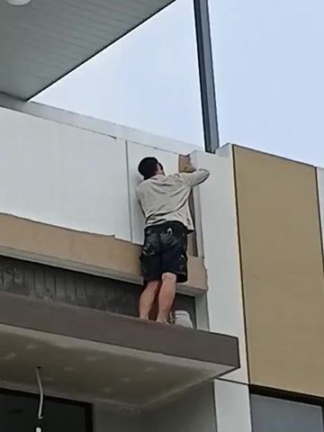 A person is standing on a ledge, painting a building exterior. The person is wearing a light-colored shirt and dark shorts. The building has white and beige panels. The sky is overcast.