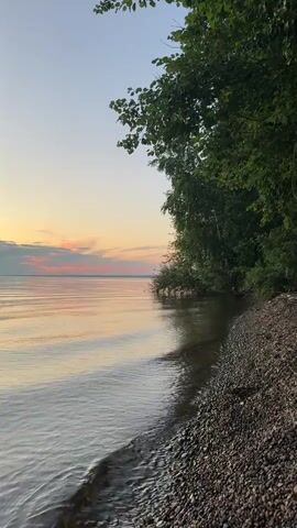 A serene video captures the tranquil shore of a body of water at sunset. The sky transitions from a pale blue to soft hues of pink and orange as the sun dips below the horizon. Gentle waves lap against a pebbly beach, lined with lush green trees that cast shadows on the water's edge. The overall atmosphere is one of peacefulness and natural beauty.