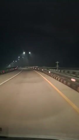 The video shows a first-person perspective from inside a vehicle driving on a multi-lane highway or bridge at night in Mukee Gogo, Indonesia. The road is well-lit by numerous streetlights positioned along the median and sides. White solid lines mark the left edge of the lane and the median, while a prominent yellow solid line runs along the right side of the current lane. Guardrails with small reflective markers line the right side of the road. The sky is dark, indicating nighttime, and no other significant features like buildings or landscapes are visible beyond the immediate road structure. Distant vehicle lights can be seen in the far background. No people or visible text are present in the frame.
