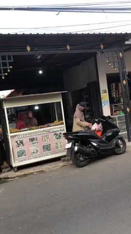 The video shows a street-level view of a food stall and a building. A woman wearing a headscarf and a beige jacket is standing next to a black scooter, holding a plastic bag. She appears to be interacting with someone inside the food stall. The food stall has a sign that reads "Spesialis Gorengan". Two women, also wearing headscarves, are visible inside the stall. There are fried foods displayed in the stall. The building behind the stall has a dark-colored roof and appears to be a shop or residence. The overall environment is an outdoor street scene.