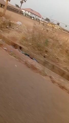 The video shows a street scene, possibly in a village or rural area. The road is reddish-brown dirt. The street edge is lined with trash and debris. Buildings appear to be old and made of simple materials like mud brick. One shot shows a man walking past the side of one of these buildings. The buildings are in a state of disrepair, with peeling paint and rusty metal shutters on the windows.  The video is shaky, suggesting it was taken while the camera was in motion, perhaps from a passing vehicle. The overall impression is one of a simple, possibly impoverished, community.