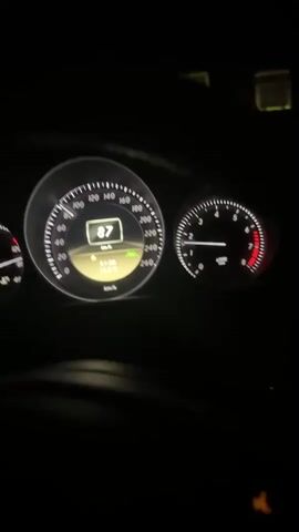 The video shows the dashboard of a car at night, with the speedometer indicating speeds between 84 and 82 km/h. The car is on a highway, as indicated by the presence of other vehicles, including a truck ahead, and traffic signs. The interior lights of the car are on, illuminating the dashboard and a digital display showing the time and temperature. The driver's hands are visible on the steering wheel. The video captures the driver's perspective as they travel down the highway.
