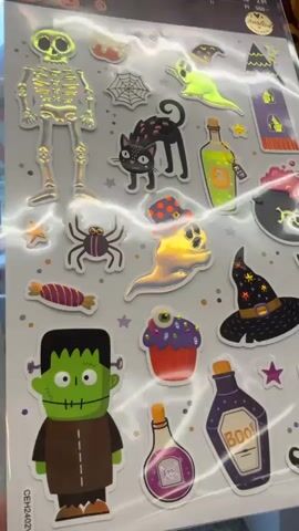 The video shows a sheet of Halloween-themed stickers on a white background. The stickers are of various Halloween characters and objects including a skeleton, a black cat, a ghost, a spider, a Frankenstein monster, witch hats, potion bottles, a cupcake with an eyeball, candy, and a haunted house. The stickers appear to be holographic or have a reflective finish, as indicated by the shimmering light reflecting off them. The video is shot from a close-up perspective, focusing on the details of the stickers. There are no people or active interactions depicted in the video, only a static display of the stickers. The environment appears to be indoors, possibly a store or a craft room, given the reflective nature of the sheet and the general lighting.