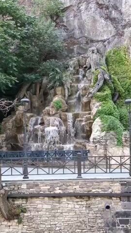 A short video clip shows a cascading waterfall that has been integrated into a rocky embankment. The waterfall is surrounded by lush green foliage and ivy, creating a naturalistic setting. In the background, a rough, grey rock face rises steeply. In front of the waterfall, a black metal fence with ornate scrollwork separates the viewer from the water feature. A paved walkway runs behind the fence. Below the walkway, there is a stone wall constructed from irregular grey and brown stones, which forms the edge of a body of water or canal. The water in the canal appears murky and shows signs of algae. The overall impression is of a carefully designed landscape feature intended to mimic a natural waterfall within an urban or semi-urban environment.
