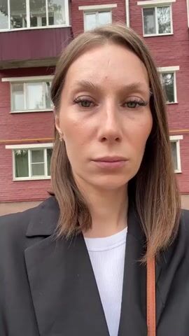 A medium shot captures a woman facing the camera, her head and shoulders filling the frame. She has medium-length brown hair that falls just past her shoulders, framing her face. She is wearing a black blazer over a white ribbed top. Her gaze is directed forward, and her expression appears neutral or somber. Behind her, a red brick building serves as the backdrop, with visible windows and balconies. The lighting suggests it is daytime. The video is short, with no apparent action or interaction taking place.