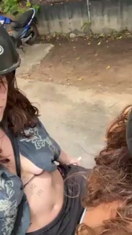 Two women are riding on a light green moped down a narrow road. The woman driving wears a helmet, a light blue tank top, and light brown pants. She has curly brown hair. The passenger wears a dark, patterned shirt over a bikini top, with black pants. She has dark hair and visible tattoos on her stomach. They are traveling down a paved road lined with trees. A black car approaches from the opposite direction. The camera appears to be held by the passenger.