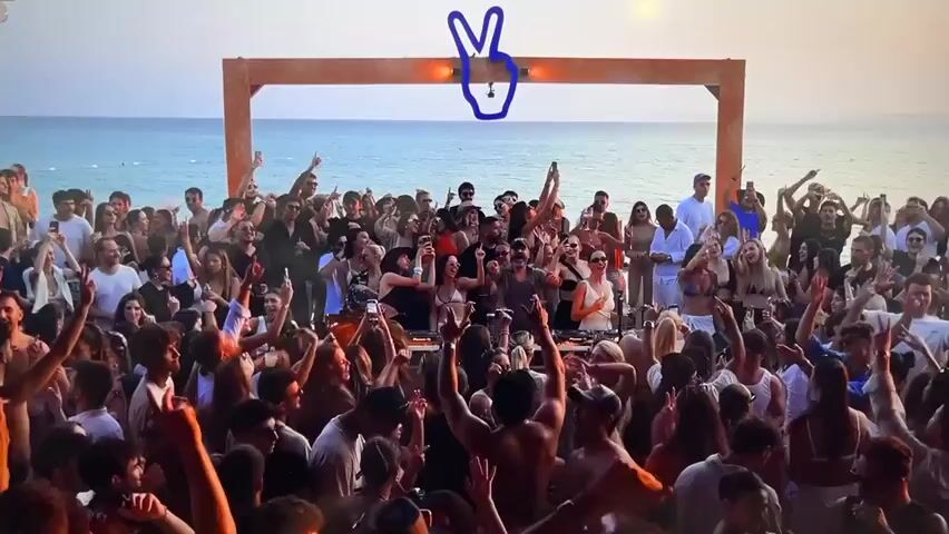 The media file shows a lively outdoor party scene on a beach at sunset. A large crowd of people, dressed in summer attire, are dancing and celebrating. Many are holding up their phones, likely capturing the moment. A DJ booth is visible in the center, with a DJ actively playing music. Above the DJ, a wooden archway is adorned with a large, stylized blue peace sign. The ocean stretches out behind the crowd, and the sky is illuminated by the warm hues of the setting sun. The atmosphere appears energetic and joyous, with individuals expressing excitement through raised arms and smiles. The time of day is clearly evening, as indicated by the sunset. While the specific location isn't explicitly stated, the beach setting suggests a coastal area. There is no visible text within the media file.