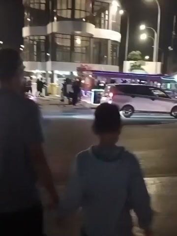 The video shows a street scene at night. In the foreground, two people, a child and an adult, are facing away from the camera, looking towards the street. A jeepney, decorated with lights, is in the middle of the street, with people on and around it. Cars are also present on the street, and a modern-looking building is visible in the background. The street appears to be wet, suggesting recent rain. The overall atmosphere is lively, with the jeepney suggesting a festive or celebratory event.