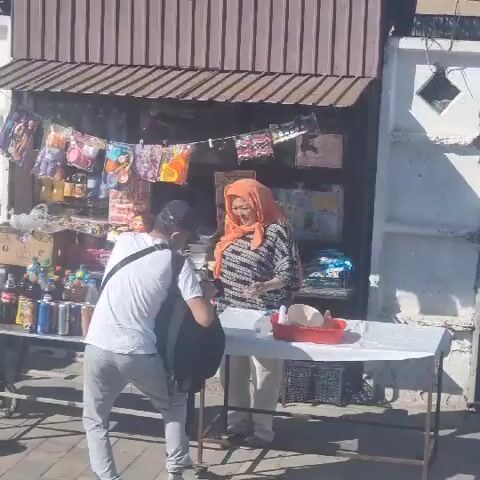 The video depicts an outdoor market scene, likely in Turkistan, Kazakhstan. The time of day appears to be sunny and warm, suggested by the strong shadows and bright lighting. The setting is a street with a small market stall that has a brown, slatted awning. The stall is laden with various goods, including drinks in bottles and cans, snacks in colorful packaging, and what appear to be jars of preserves or sauces.

In the foreground, a person wearing a white t-shirt and grey pants is seen from behind, holding a black backpack. They are interacting with a woman at the market stall who is wearing an orange headscarf and a patterned shirt. The woman is facing the person and appears to be showing them something on the table. On the table in front of the woman, there's a red plastic container with some round, light-colored objects inside, possibly food items like cheese or bread.

Further back, another stall is visible, with two women attending to customers. One woman is wearing a green dress and a headscarf, while the other is wearing a red apron over a patterned dress and a pink headscarf. They are interacting with another customer who is partially visible. This stall also displays a variety of produce, including what looks like bags of apples and watermelons in crates.

A sign with text in Cyrillic and English is visible on a building behind the stalls. The Cyrillic text reads "ДУКЕН МАГАЗИН" and the English text reads "SHOP". This confirms the location is a shop or market area. The overall impression is a lively street market scene on a pleasant day.