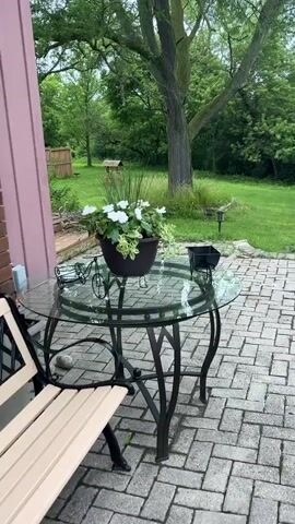 The video shows an outdoor patio area with a glass-top table and chairs. A potted plant with white flowers sits on the table. Beyond the patio is a grassy backyard with a wooden fence, a tree, and a small well. The scene appears to be peaceful and uneventful.