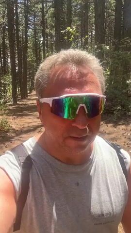 A man wearing a grey t-shirt and white, rainbow-lensed sunglasses is walking on a dirt path through a forest. The man is looking at the camera and talking. He is wearing a black backpack. The trees are tall and dense, and the ground is covered in dirt and pine needles. Sunlight filters through the trees, creating dappled light and shadows.