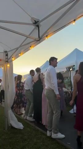 The video displays an outdoor evening event under a series of white canopy tents adorned with string lights. Approximately 10 to 15 adults are visible, most dressed in semi-formal attire. A man in a white long-sleeve shirt and khaki pants stands on a dark-colored platform with his back partially to the camera, swaying to music. To his right, a woman in a maroon midi dress is actively dancing, turning and gesturing with her arms. Other individuals are scattered behind them, including women in floral, black, and light pink dresses, and men in white shirts and various colored pants. A man in a grey collared shirt and dark trousers walks across the frame on the right side. Briefly, a man wearing a kilt and a green vest is visible in the background, raising his arms. The ground beneath the tents appears to be grass. The camera pans slightly right and then back left, capturing different angles of the dancing and mingling guests.