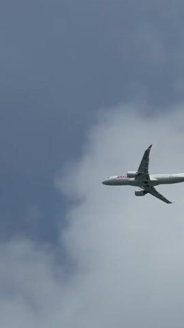 A wide shot, low angle video shows a white airplane with red markings flying through a cloudy blue sky. The airplane is seen from the side as it moves from left to right across the frame. The tail of the airplane has a red cross on a white background, the Swiss flag. The body of the airplane has red lettering that appears to say "SWISS". The registration number on the tail is "HB-JCT". There are some wispy white clouds in the sky. The bottom of the frame shows a blurry, grey, curved object, possibly the top of a building or structure.