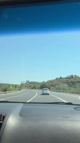 The video is filmed from inside a car, showing the view through the windshield and side windows. The car is driving on a highway on a clear, sunny day. The highway has multiple lanes and is surrounded by green vegetation and rolling hills. A white car is visible in front of the camera car, and there are road signs and markers along the side of the road. The side mirror reflects the passenger taking the video. There are stickers on the windshield, one of which appears to be a safety sticker for the airbag.