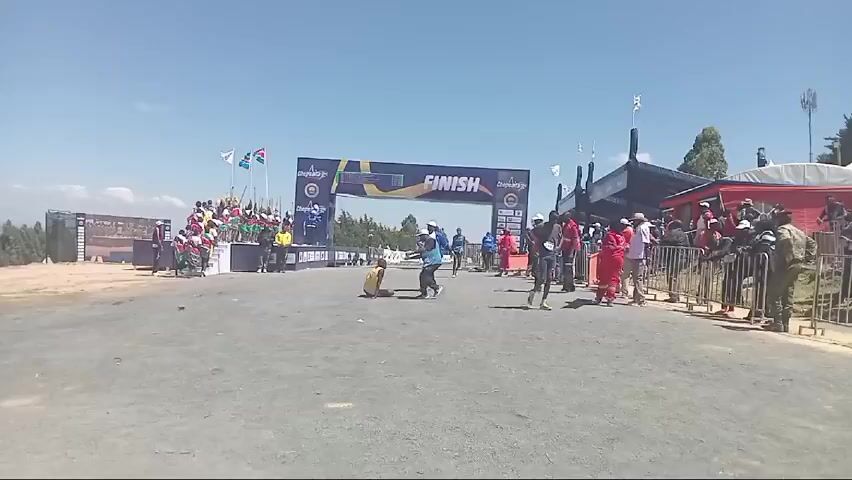 The video shows the finish line of a marathon on a sunny day. A large banner that says "FINISH" is positioned across the road. Runners are approaching the finish line, some are walking or being assisted by staff. People are standing behind a metal barrier on the right side of the road, including security personnel in military uniforms. On the left side of the road, there is a platform with people dressed in colorful traditional clothing, waving spears. A scoreboard above the "FINISH" banner displays the race times.  An emergency medical worker is carrying a stretcher. Several flags are visible, including what appears to be the flags of Kenya, Uganda, and South Sudan. The ground is a gray, gravelly surface. The camera movement is unsteady, and there is a brief moment where the view is obstructed.