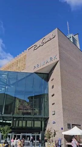 The video shows an exterior view of a large building with glass windows and a sign that reads "Primark". It is a sunny day with a blue sky and some clouds. There are people walking in front of the building and several cars parked nearby. The footage is shot from a slightly low angle, looking up at the building. It appears to be a commercial area with a market set up in front of the buildings.