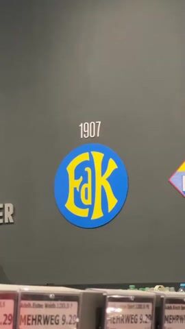 The video pans across a dark gray wall displaying historical logos and dates. At the beginning, the year 1907 is visible, followed by a blue circular logo with yellow letters "FAK". The camera then moves to show the year 1911 above a diamond-shaped logo that reads "EDEKA" in yellow lettering on a background of yellow, blue, and pink. Finally, the year 1912 is displayed next to a rectangular logo that also says "Edeka" in yellow lettering on a blue background. The bottom of the frame shows a row of various products in bottles and packaging on shelves, including sodas and juices.