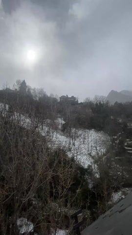 The video depicts a landscape with light snowfall. In the foreground, bare trees and bushes are visible. Patches of snow cover the ground. In the midground, a building with a pointed tower stands on a hill, along with other buildings and more trees. In the background, mountains are faintly visible through the overcast sky. The sun is obscured by clouds, creating a diffused light. Snowflakes fall throughout the scene. The camera appears to be stationary.