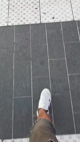 The video begins with a close-up shot of a person's white sneaker stepping onto a textured, raised tactile paving surface. The camera then pans up and to the left, revealing a street scene with a building on the right. The building has signs that read "NICEA IMMOBILIER" and "NICEA PROMENADE". Trees are in the foreground, and a streetcar track is visible. In the background, there are more buildings, palm trees, and the ocean. The sun appears to be setting, casting a warm glow over the scene. Cars are seen driving on the street, and a bicycle is parked near the building. The video ends as the sun is partially obscured by trees, creating a silhouette effect.