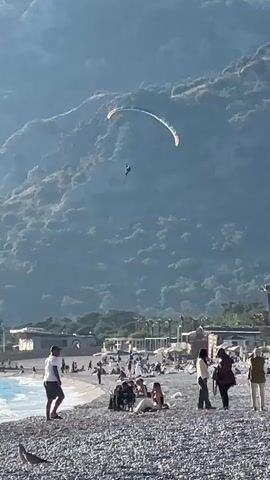 The video shows a paraglider coming in to land on a rocky beach. The paraglider is white with green, orange and red stripes, and has the word "PILOTS" written on it. Below, people are walking around the beach. A couple walks away from the camera, the man wearing a hat and blue backpack. In the background is a large hill covered in trees. The sky is clear and blue. The beach is made up of small stones. There are a few palm trees and some buildings in the background. Some people are sitting, others are standing and looking. The video is taken in daylight.