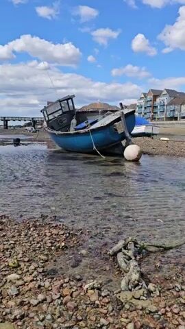 A weathered blue boat is anchored in shallow, clear water on a rocky shore. The boat is tilted, with its bow dipping into the water and its stern resting on the exposed seabed. A white buoy floats alongside the stern. In the background, a pier extends into the water, and buildings with multi-colored facades line the shore. The sky is blue with scattered white clouds. A thick, knotted rope lies on the rocks in the foreground.