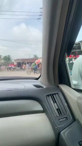 The video, captured from inside a car, shows a street scene from the driver's perspective. The car is stationary or moving very slowly at an intersection or busy street. The front dashboard is visible in the foreground. Through the windshield, a bustling street is seen with multiple motorbikes, some carrying passengers, and pedestrians moving around. There are market stalls or small shops along the roadside with awnings, and several people are gathered around these establishments. Utility poles with numerous wires crisscross the sky, suggesting an urban or semi-urban environment. The sky is overcast, indicating a cloudy day.